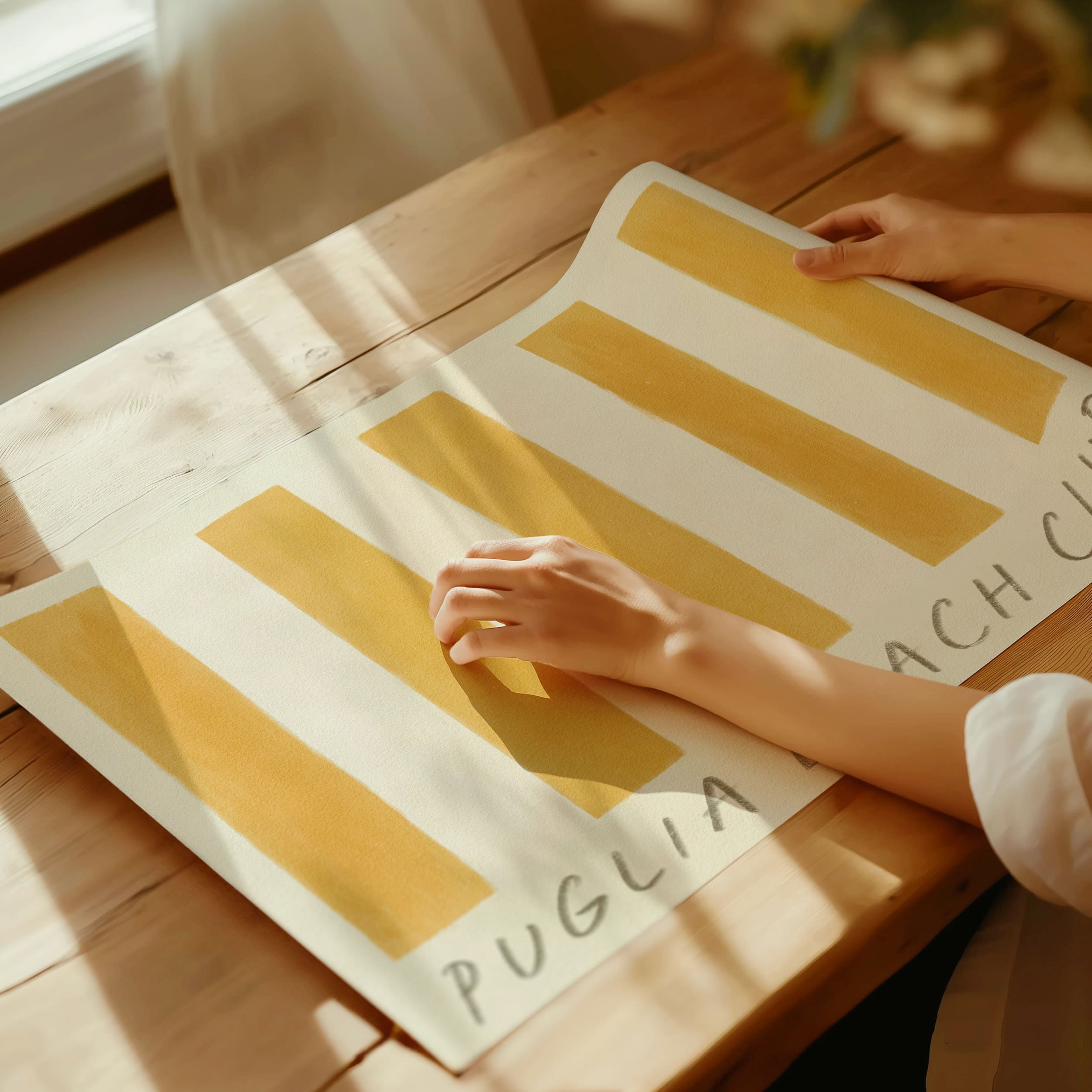 Puglia beach club stripe giclee art print of original artist oil painting in a rich deep ochre yellow. This artwork by Cornwall artist Mads is a replica of a yellow retro stripe beach towel. In this photograph, the art print has been un rolled in a sun lit room and shows the hands of a woman. Sun light shadows dapple across the oil art print. Art prints come in a cylinder postage tube for maximum protection in the post.