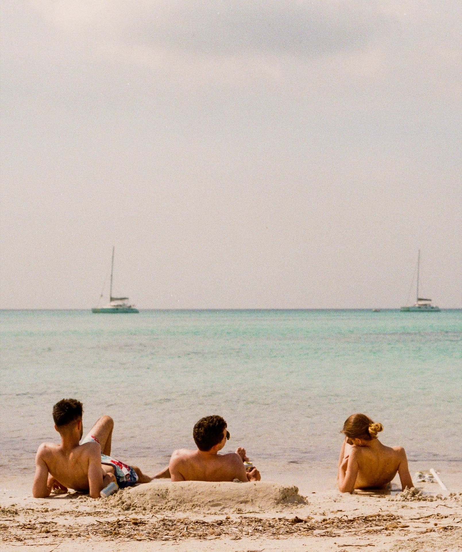 friends sunbathing by salty blue european sea with sail boats and fluffy white clouds
