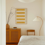 Puglia beach club stripe giclee art print of original artist oil painting in a rich deep ochre yellow. This artwork by Falmouth artist Mads is a replica of a retro stripe beach towel. In this photograph, the cotton paper puglia beach art print hangs in a thin oak frame on off-white painted bedroom wall. Paint colour by Atelier Ellis. The art is displayed in a modern bedroom with European styling and Scandinavian accents.