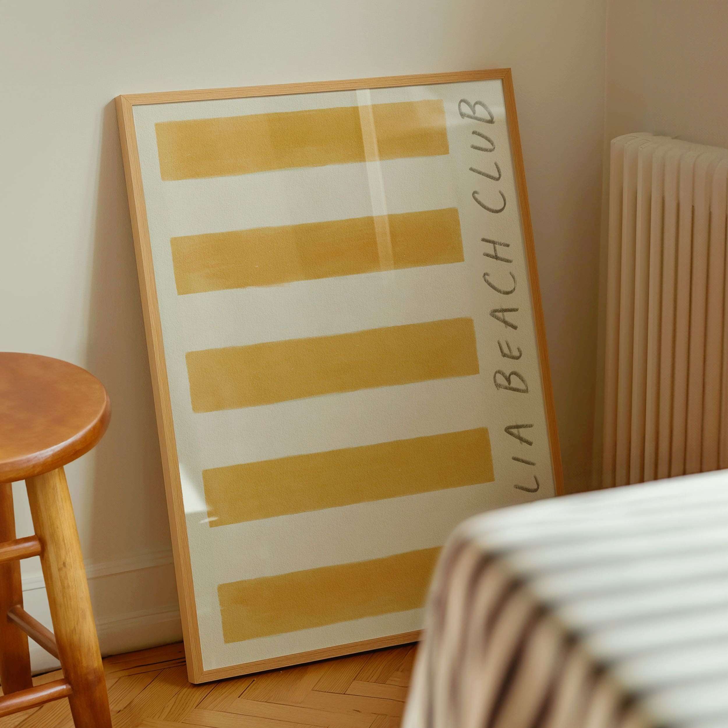 puglia beach club stripe giclee art print of original artist oil painting in a rich deep ochre yellow. art print leans in a thin pine wood glass frame against farrow and ball white interior painted walls on traditional french parquet wood floor tiles. a stripe tablecloth drapes dinning room table and a traditional vintage wooden stall appears to the left of the frame. a victorian cream colour radiator hangs on the wall to the right