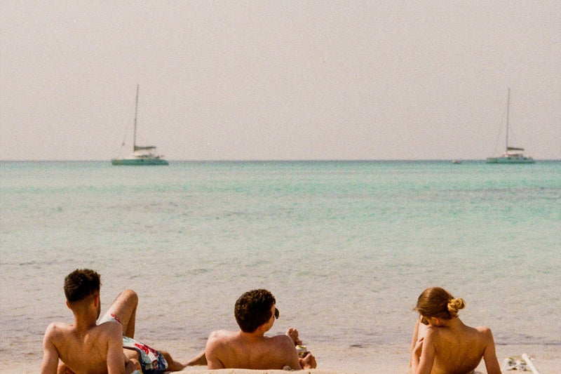 friends sunbathing by salty blue european sea with sail boats and fluffy white clouds
