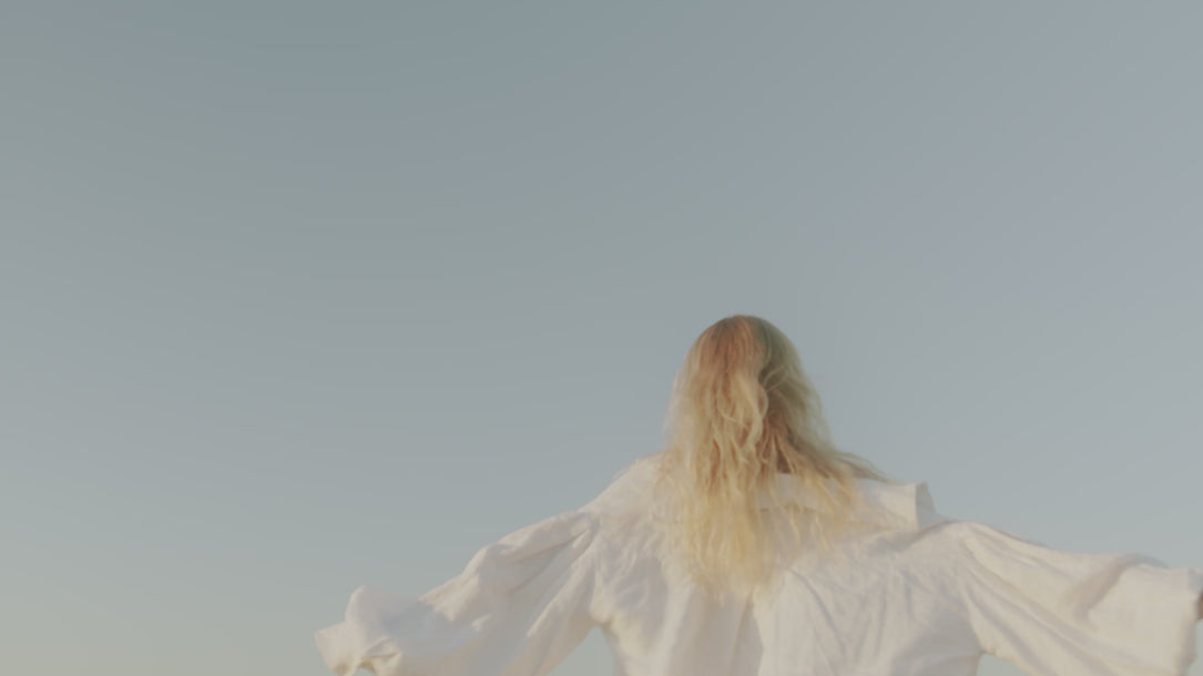 madsunsea blonde hair whimsically waving to plane in blue sky wearing white linens